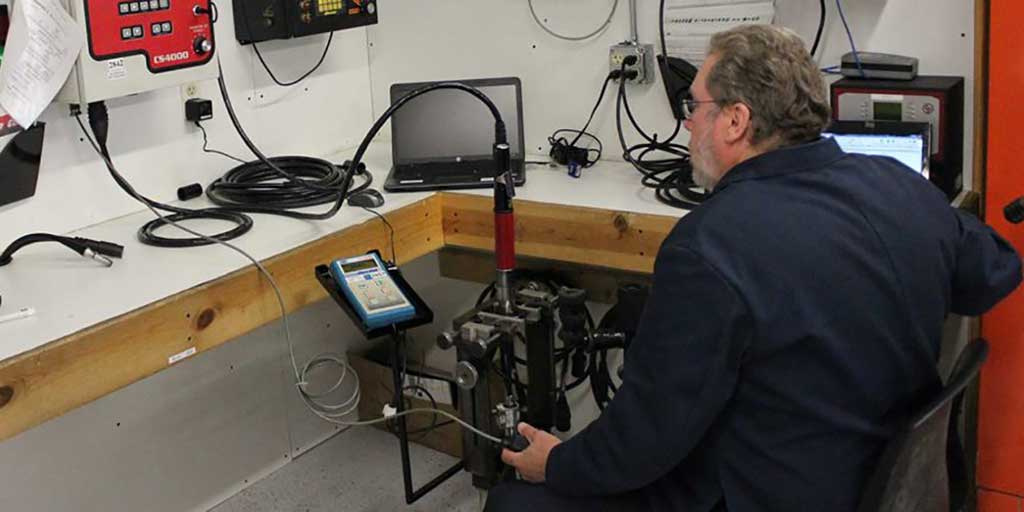 Technician using diagnostic equipment in a workshop, surrounded by meters and cables.